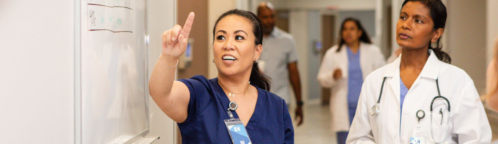 Young adult female nurse gestures towards the whiteboard while meeting with her team